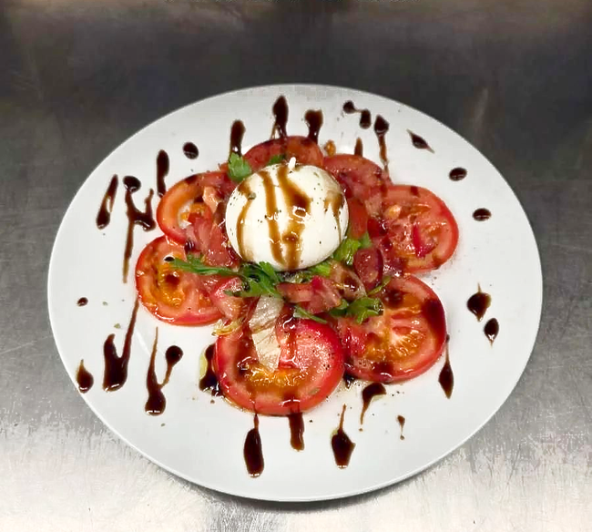 Burrata sur des tranches de tomates et des feuilles de menthe, le tout arrosé d'un filet de glaçage balsamique et présenté sur une assiette blanche.