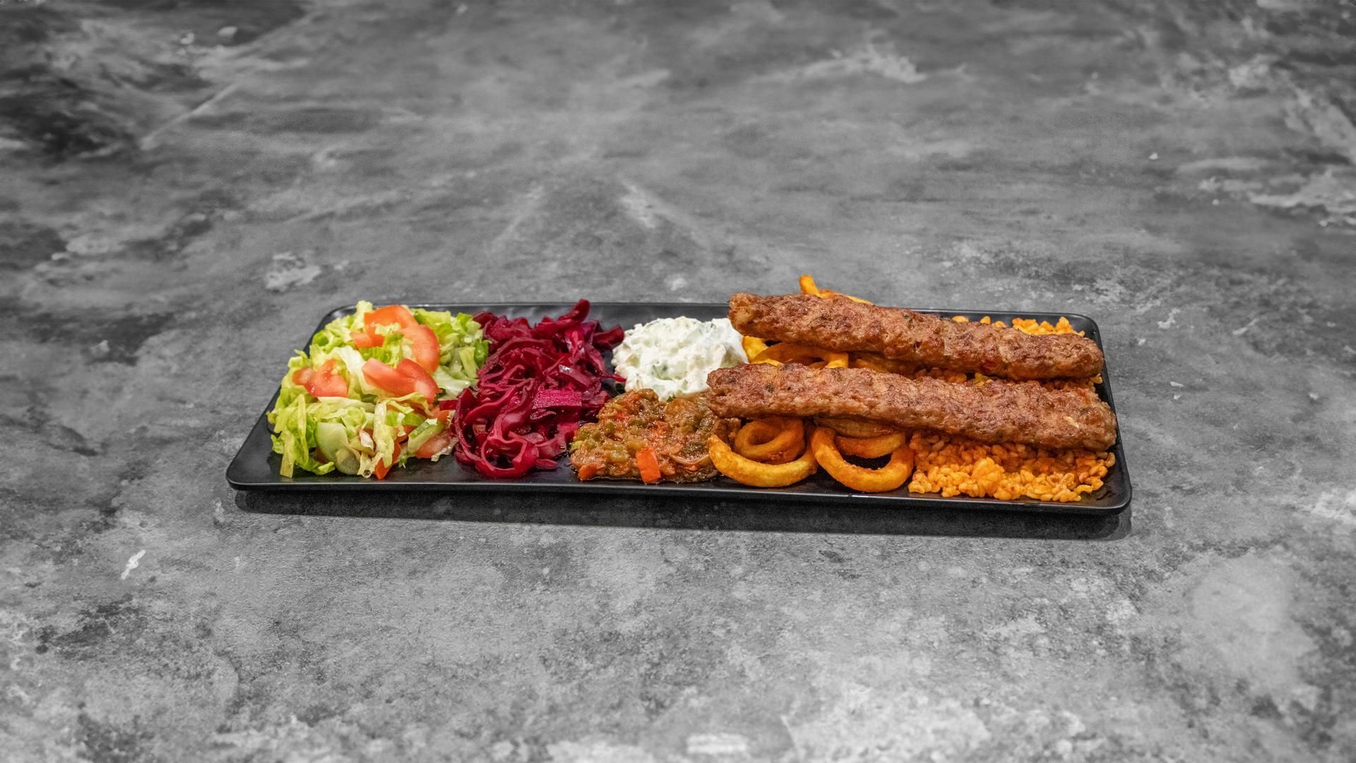 Une assiette rectangulaire de spécialités turques comprenant deux brochettes de kebab, du riz, des rondelles d'oignon frit, du chou rouge et une salade d'accompagnement.