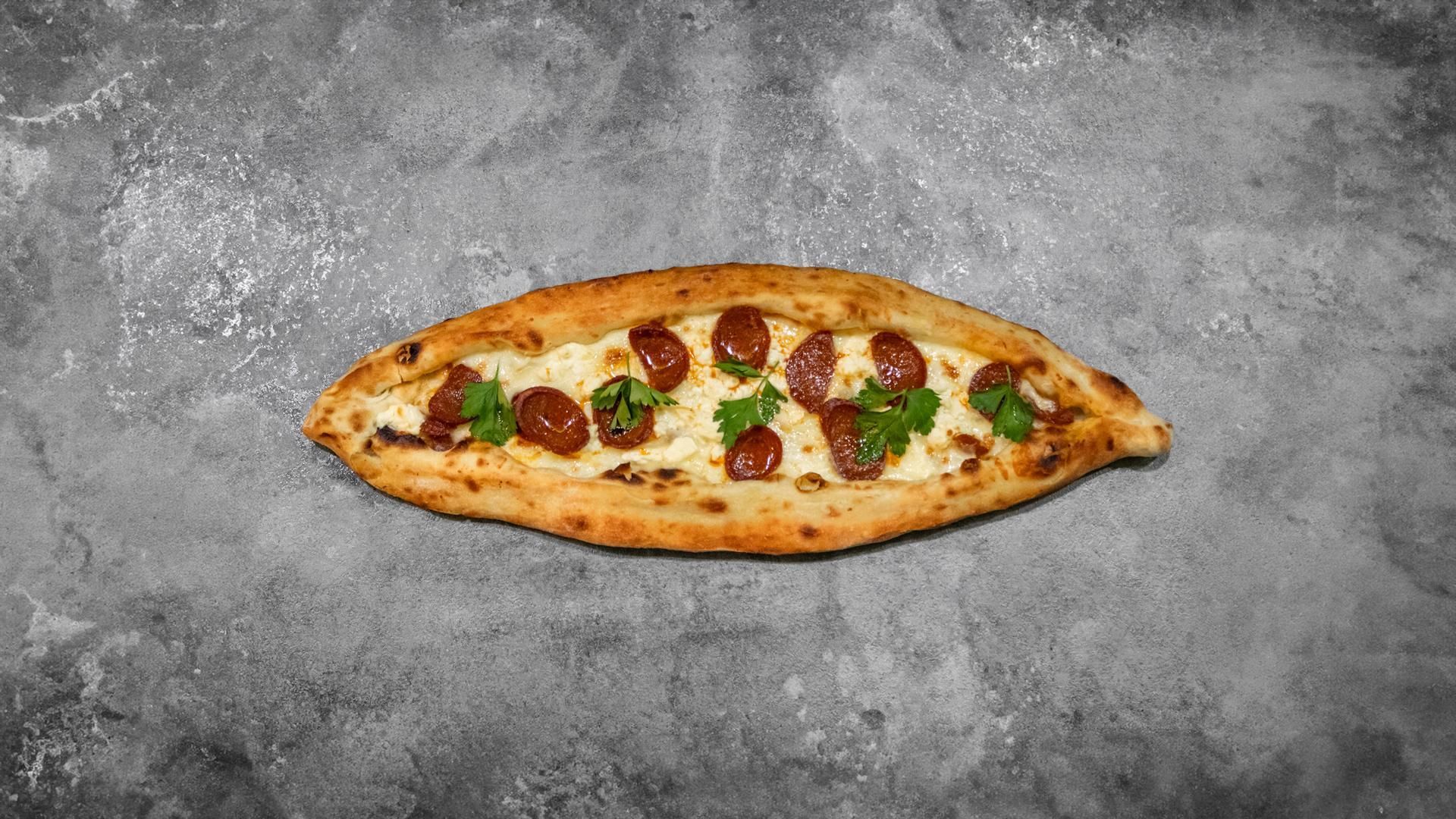 Une galette en forme de bateau garnie de fromage fondu, de tranches de chorizo et d'herbes fraîches, sur une surface en pierre grise.