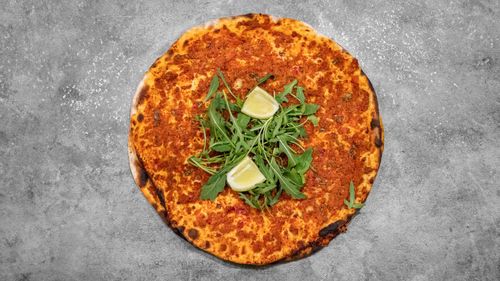 Un lahmacun turc garni de roquette fraîche et de deux quartiers de citron sur un fond gris texturé.