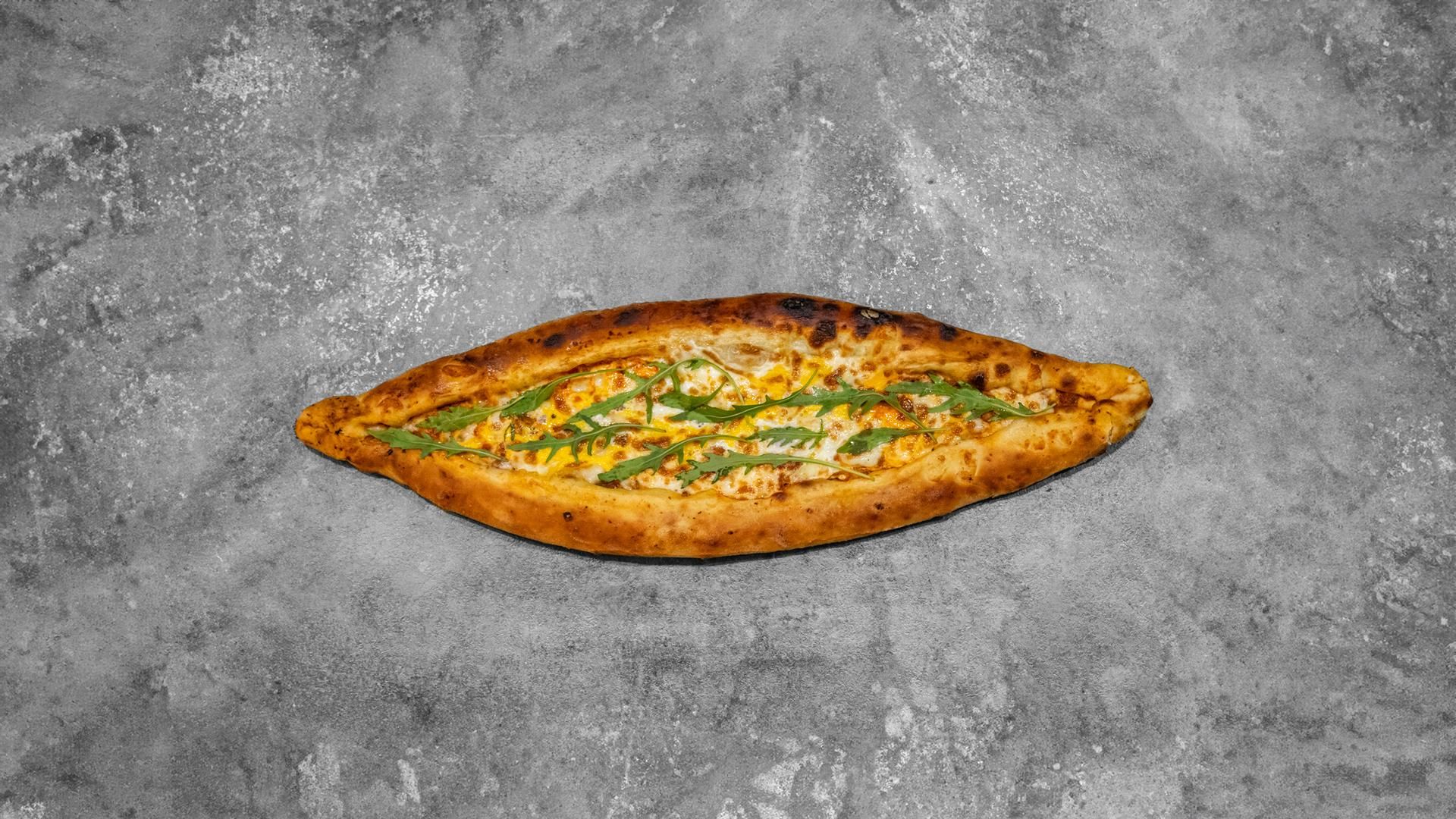 Un pain plat turc pide en forme de bateau, garni de fromage fondu et d'herbes vertes, sur un fond gris texturé.