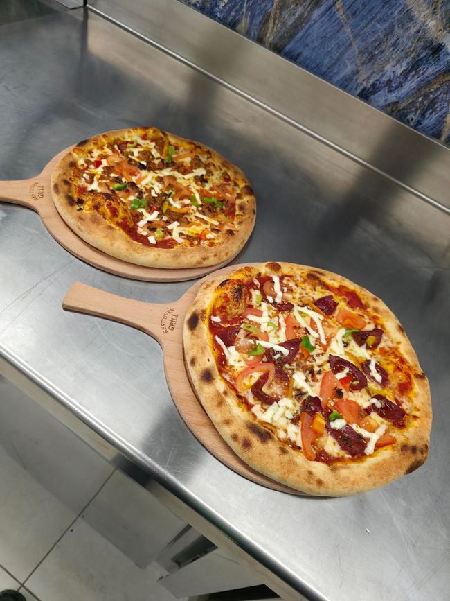 Deux pizzas fraîchement cuites avec des garnitures variées, servies sur des pelles à pizza en bois sur un comptoir en acier inoxydable.