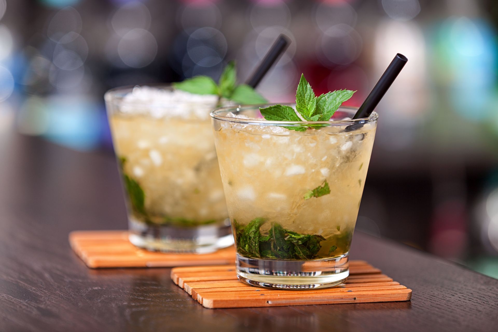 Deux verres de cocktails garnis de menthe, avec des pailles noires, posés sur des sous-verres en bois sur le comptoir d'un bar.