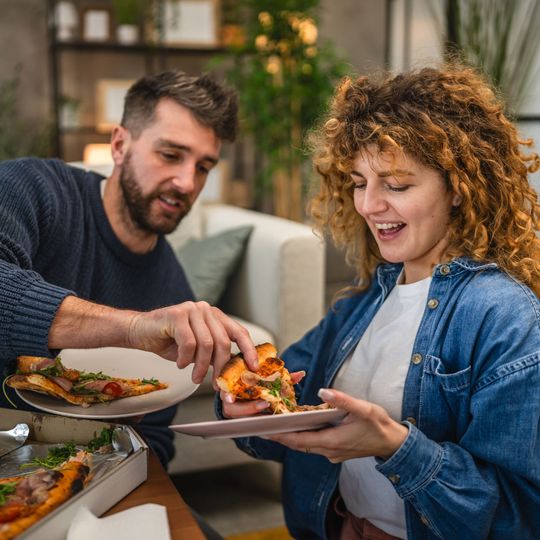 Un homme et une femme sourient en servant des parts de pizza dans des assiettes, dans un cadre intérieur chaleureux.