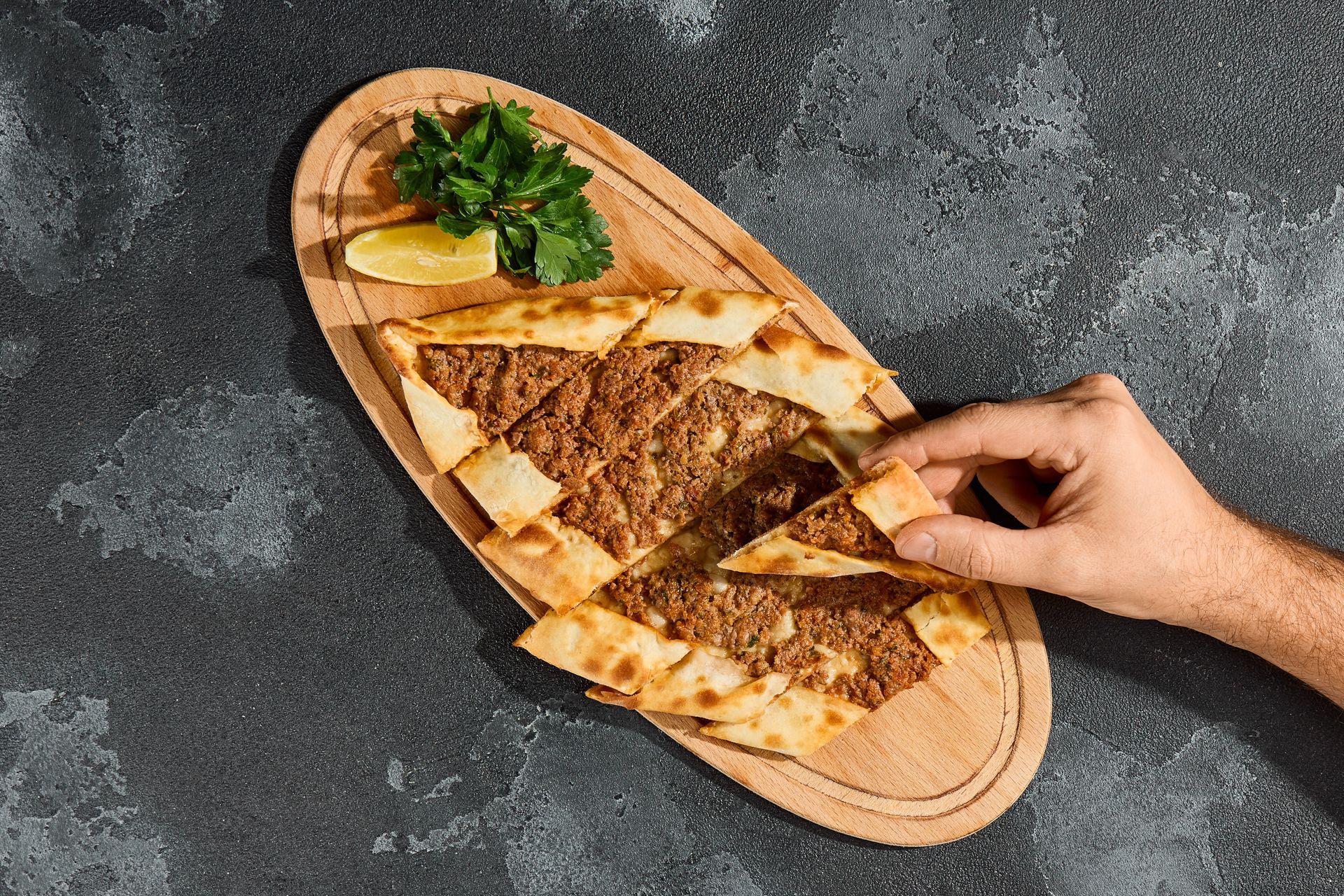 Une main saisit une tranche de pide turque garnie de viande assaisonnée sur une planche en bois, à côté d'un bouquet de persil et d'un quartier de citron.