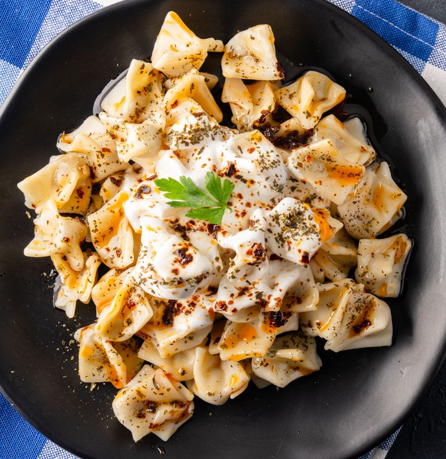Une assiette noire de mantis (raviolis turcs) assaisonnés de yaourt, d'un filet d'huile pimentée, de menthe séchée et d'un brin de persil.