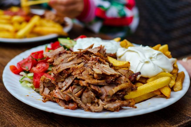 Une assiette de viande de gyros servie avec des frites, une salade de tomates et de concombres frais, et deux quenelles de sauce blanche crémeuse.