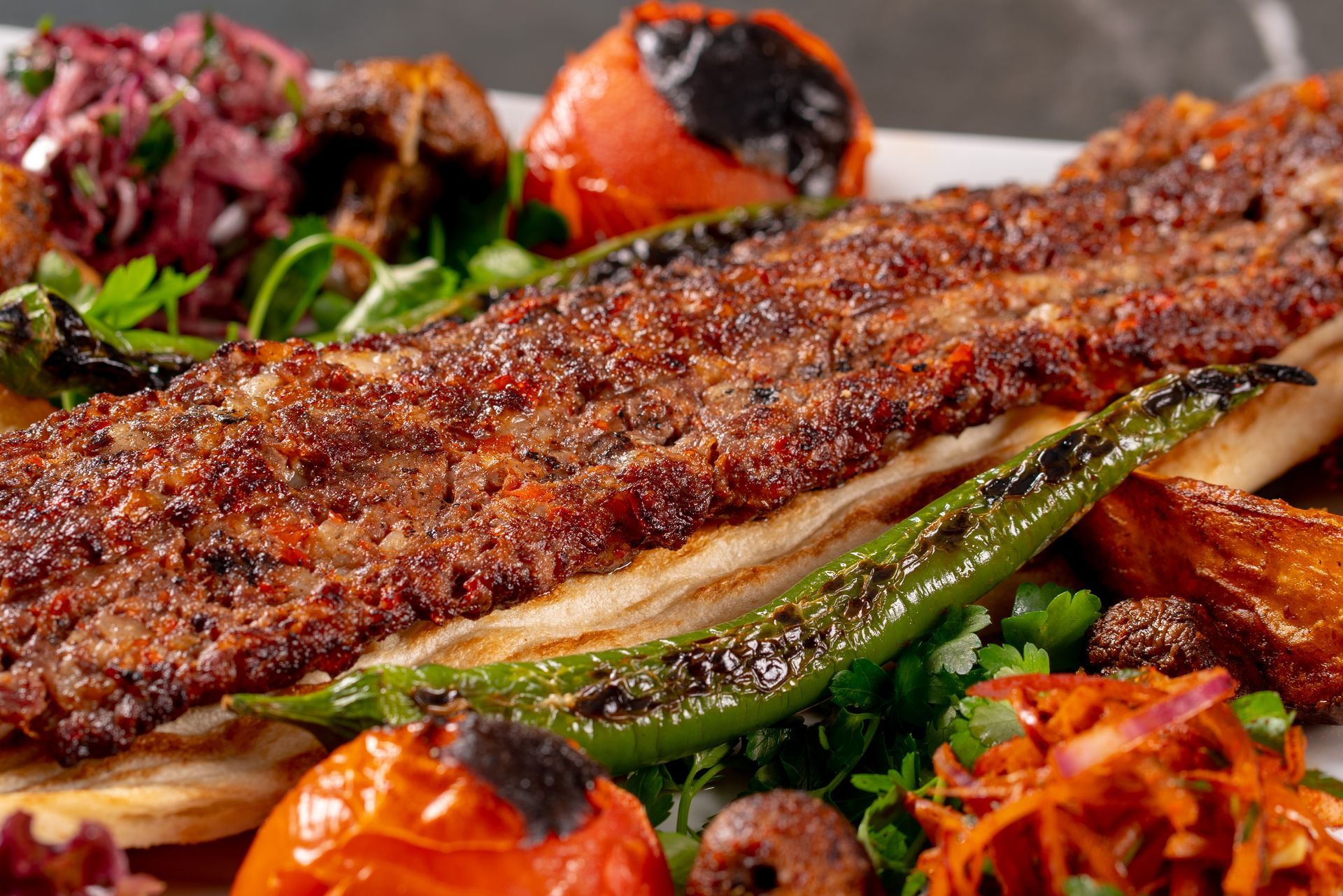 Une longue brochette de viande grillée servie sur du pain plat, accompagnée de tomates rôties, d'un piment vert et de salades fraîches.