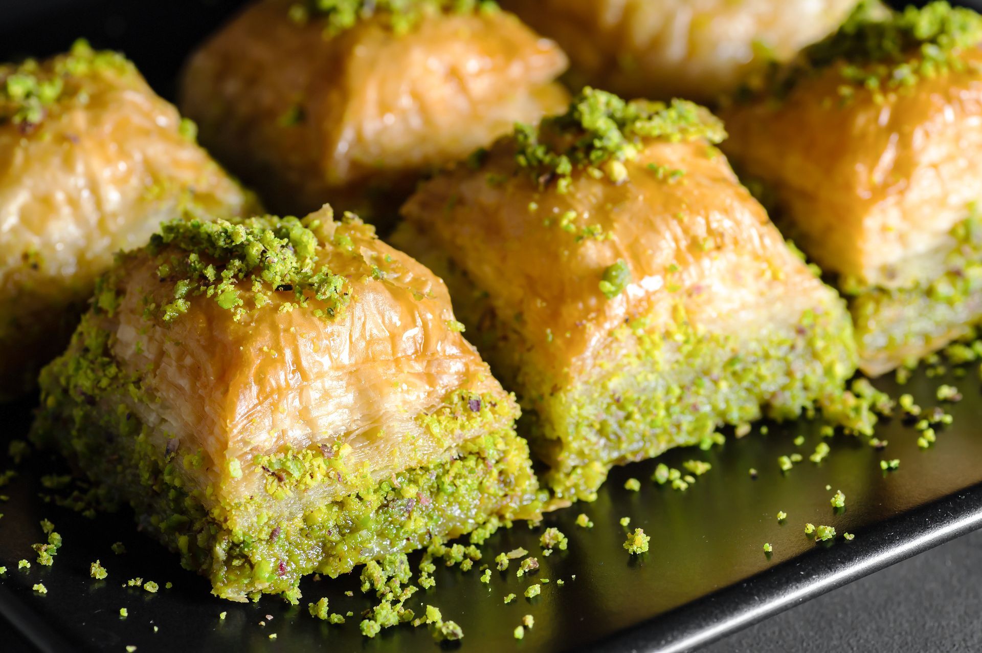 Gros plan sur plusieurs carrés de baklava doré et feuilleté, recouverts de pistaches vertes concassées, présentés sur une assiette noire.