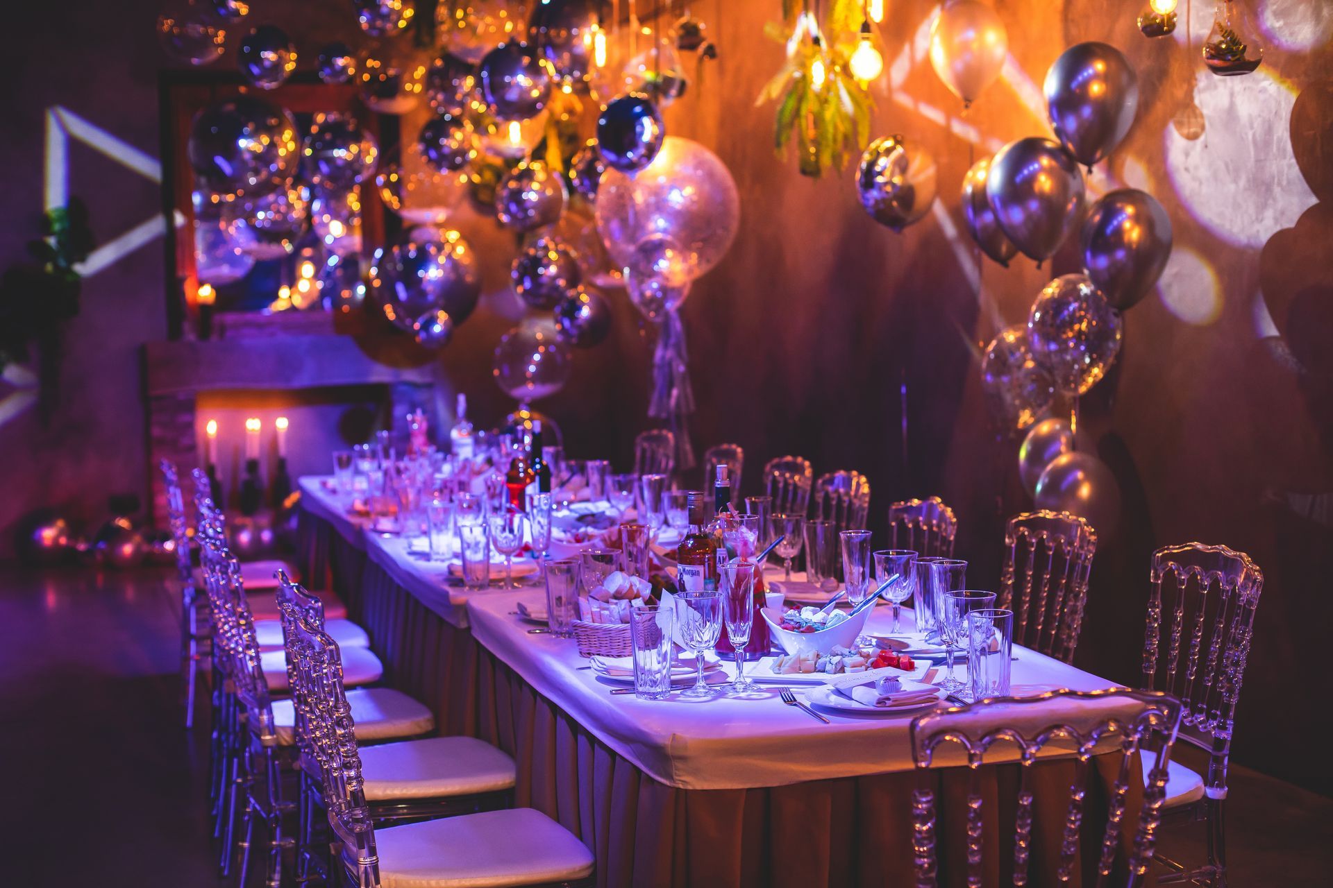 Une table dressée pour un dîner formel, ornée de nombreux ballons argentés, de bougies scintillantes et de chaises transparentes.