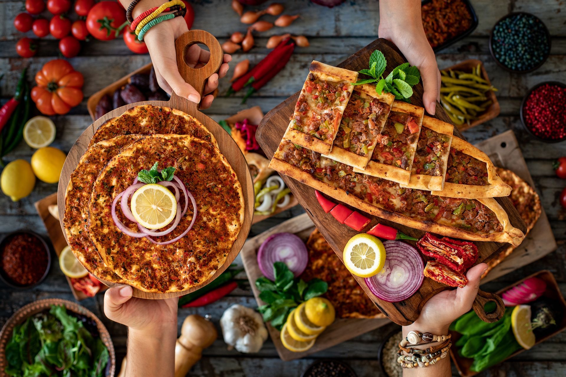 Deux mains tiennent des planches en bois présentant des lahmacuns et des pides turcs, entourés de légumes frais et d'herbes aromatiques éparpillés.