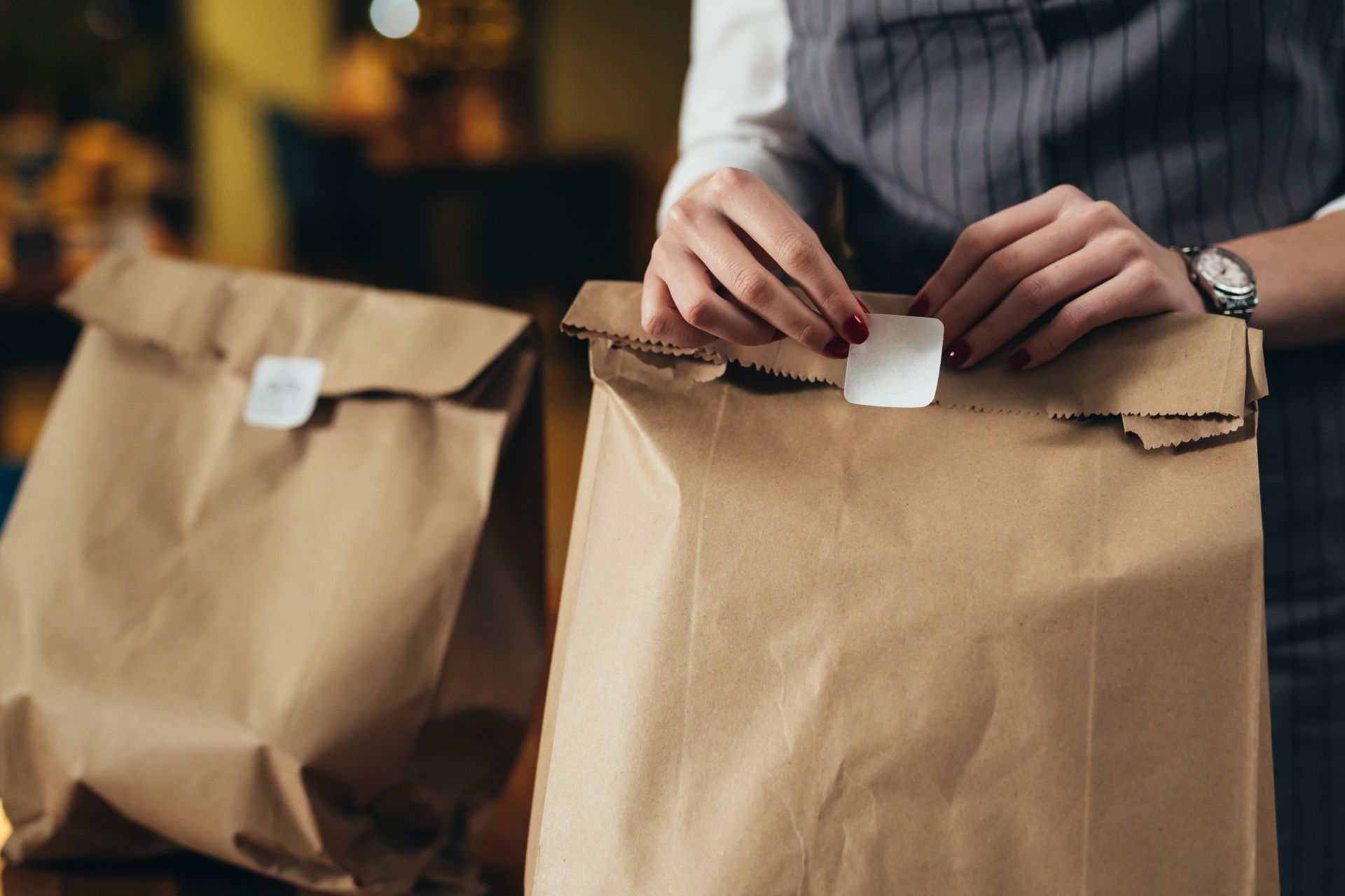 Dans un restaurant, une personne portant un tablier gris scelle un sac à emporter en papier brun avec une étiquette adhésive blanche.