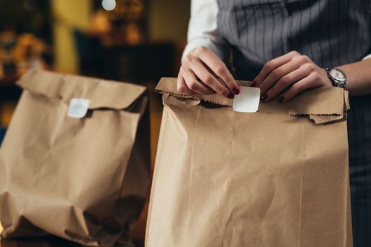Dans un restaurant, une personne portant un tablier gris scelle un sac à emporter en papier brun avec une étiquette adhésive blanche.