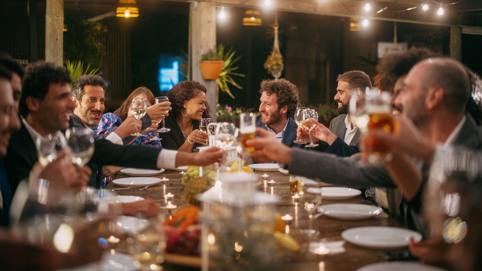 Un groupe d'amis aux profils variés trinquent lors d'un dîner en plein air, sous une lumière d'ambiance chaleureuse.