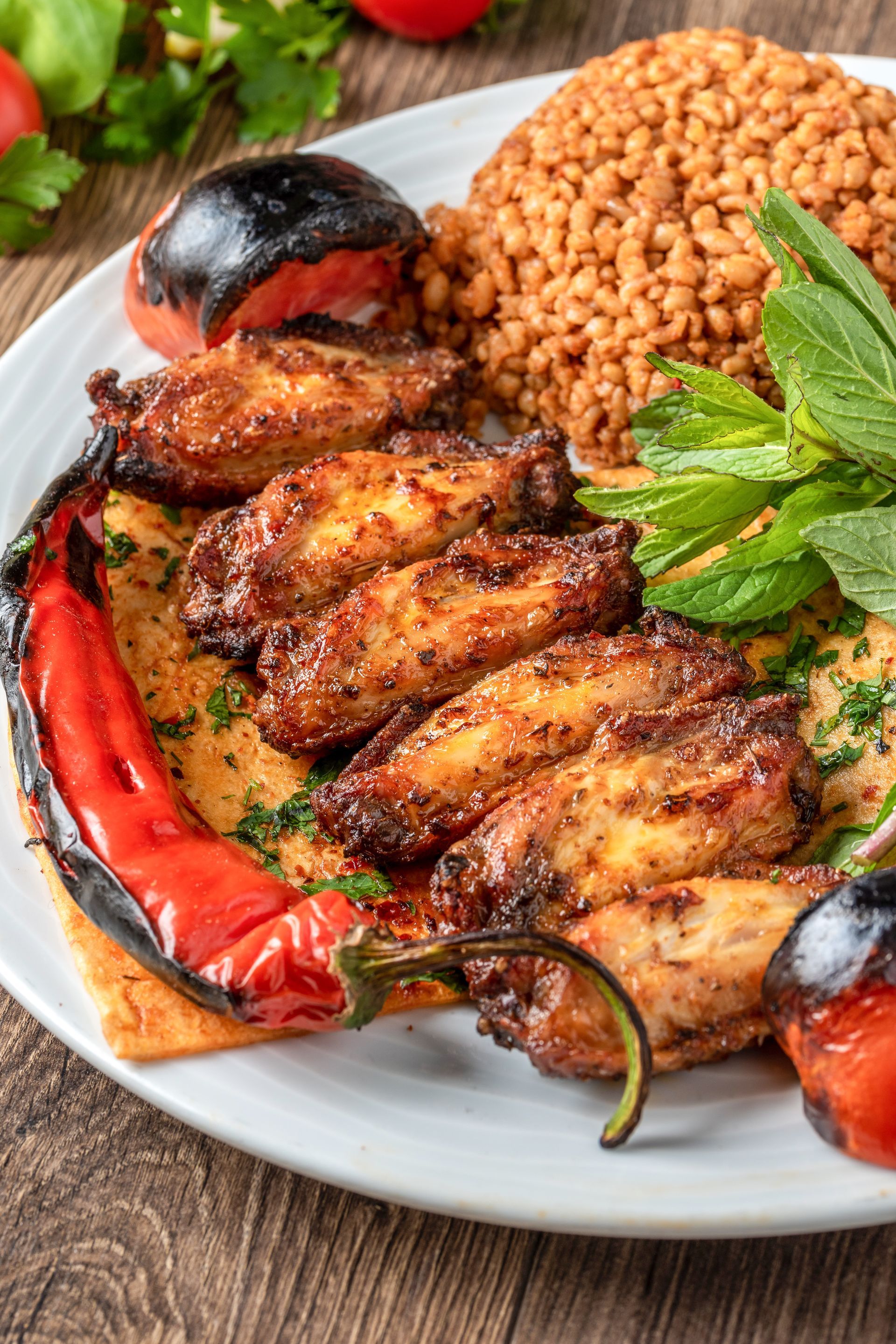 Une assiette d'ailes de poulet grillées, de pilaf de boulgour, de poivrons rouges grillés et de tomates, servie sur du pain plat avec des herbes fraîches.