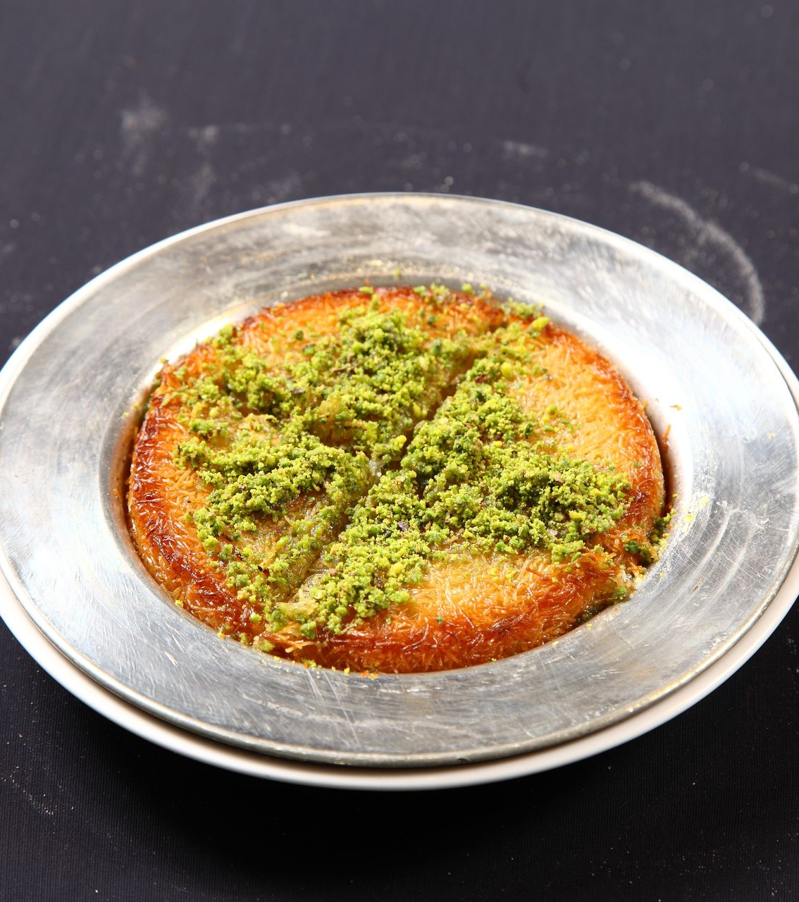 Une pâtisserie künefe ronde et dorée, garnie de pistaches vertes concassées, servie sur une assiette ronde en métal.