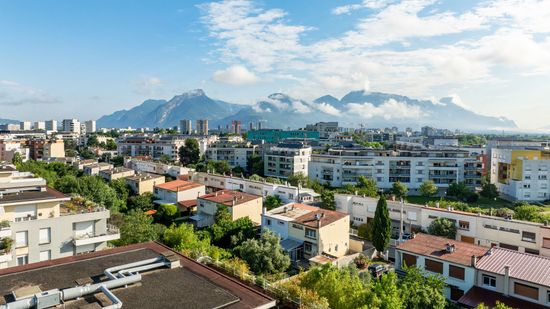 Un paysage urbain ensoleillé, composé de bâtiments bas et d'arbres, se détachant sur un fond de montagnes majestueuses partiellement nuageuses.