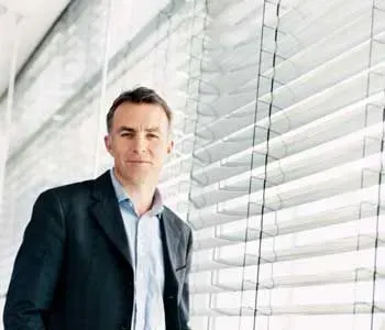 Um homem de terno está parado em frente a uma janela com persianas.