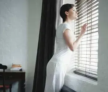 Uma mulher de vestido branco está olhando pela janela com persianas.