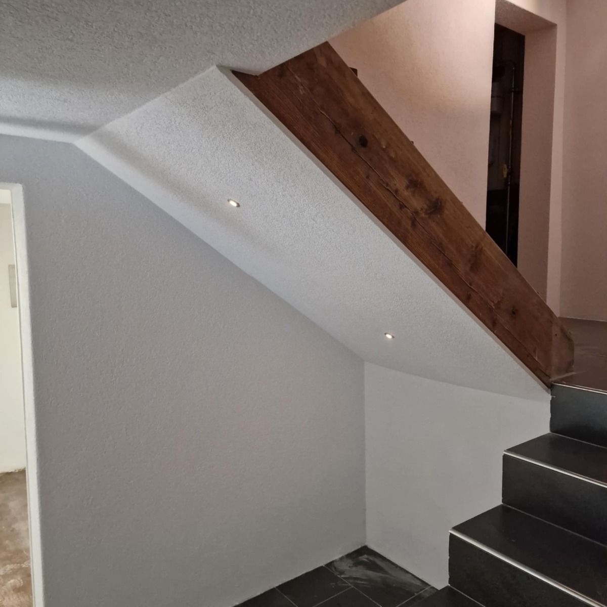 Treppe mit schwarzen Stufen, weißen Wänden und einem seitlichen Holzbalken. Einbauleuchten erhellen den Raum.