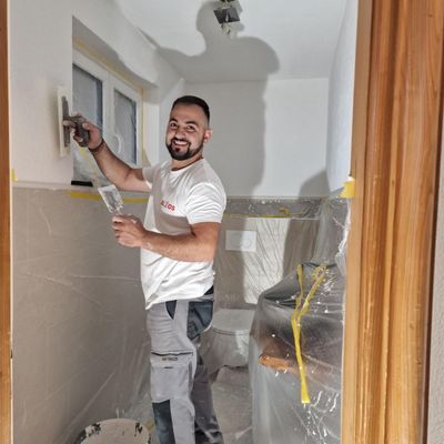 Mann streicht mit einem Pinsel eine Wand; trägt ein weißes T-Shirt, graue Hosen und einen Eimer Farbe.