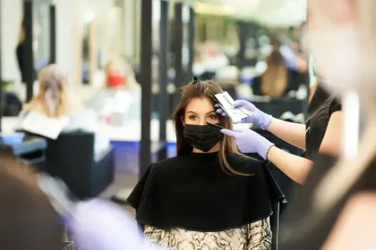Una estilista con guantes azules trabaja en el cabello de una clienta en una peluquería; ambas llevan mascarillas negras.