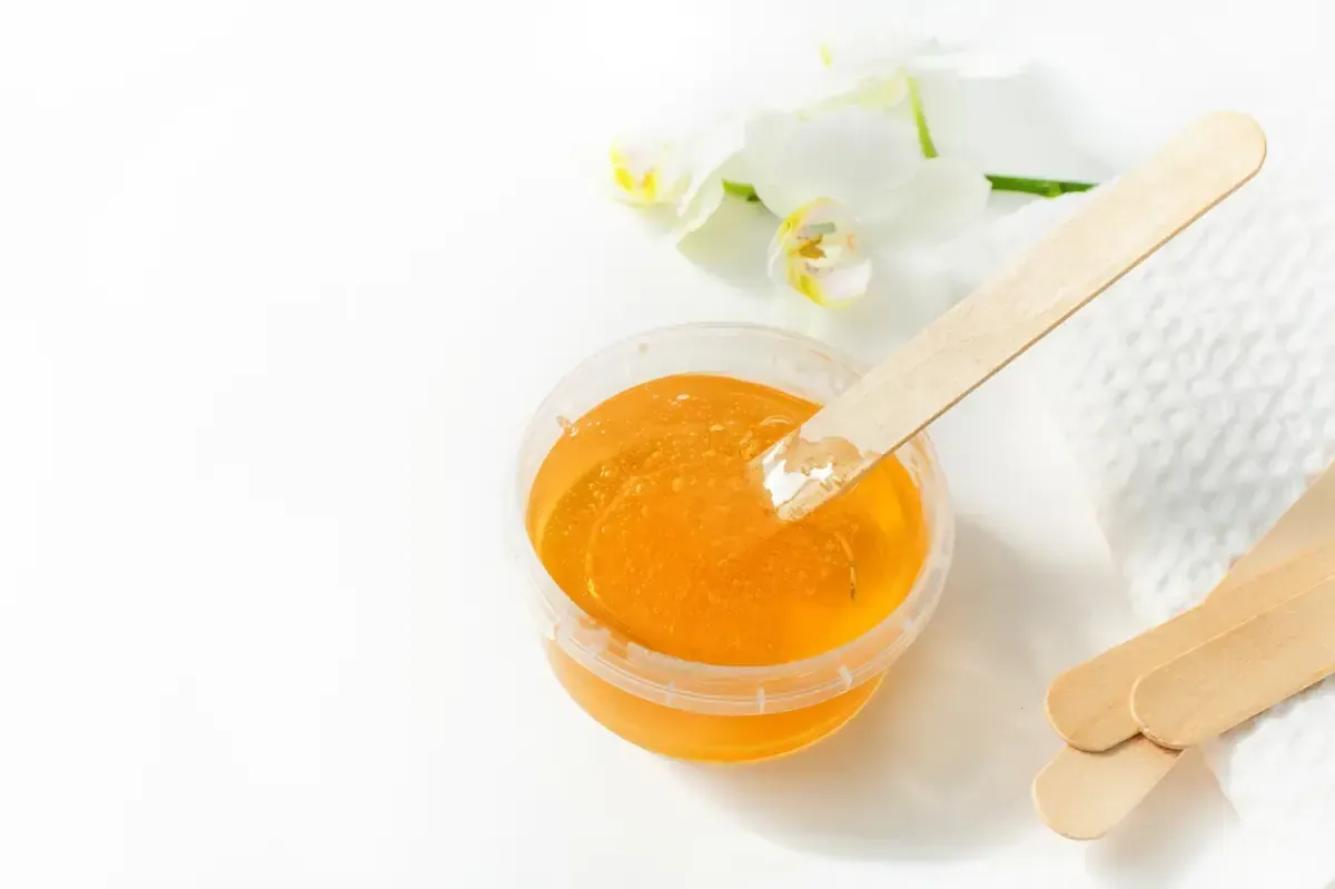 Recipiente para cera con aplicador de madera, pañuelo de papel y orquídea blanca sobre superficie blanca.