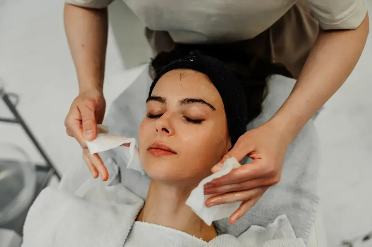 Persona recibiendo tratamiento facial, manos sosteniendo toallitas para limpiar la cara, entorno de spa.