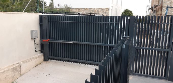 Una puerta de metal oscuro con barras verticales y abridor automático que conduce a un camino de entrada.