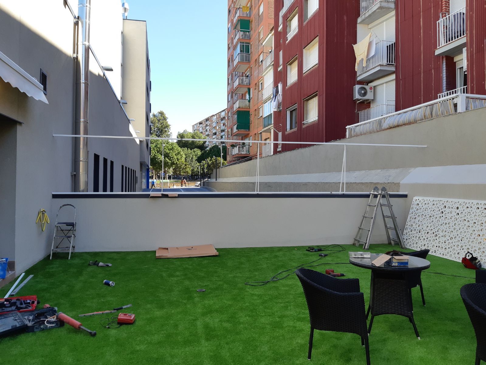 Un patio trasero con césped artificial, una valla decorativa blanca y herramientas de construcción. Al fondo, edificios de apartamentos marrones y rojos.