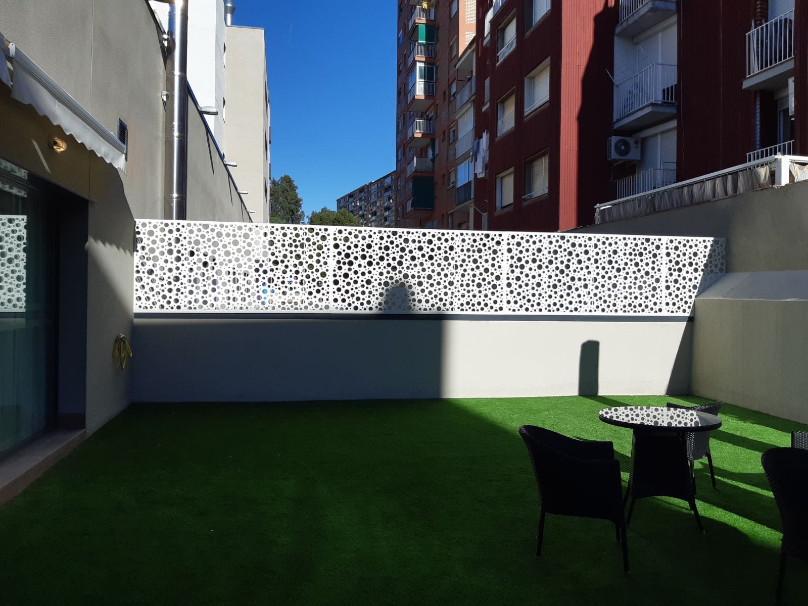 Patio trasero con césped verde, cerca blanca estampada y una mesa con sillas. Edificios al fondo bajo un cielo azul.
