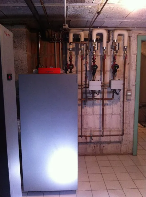 Local technique au sous-sol avec système de chauffage et tuyauterie sur mur en béton.