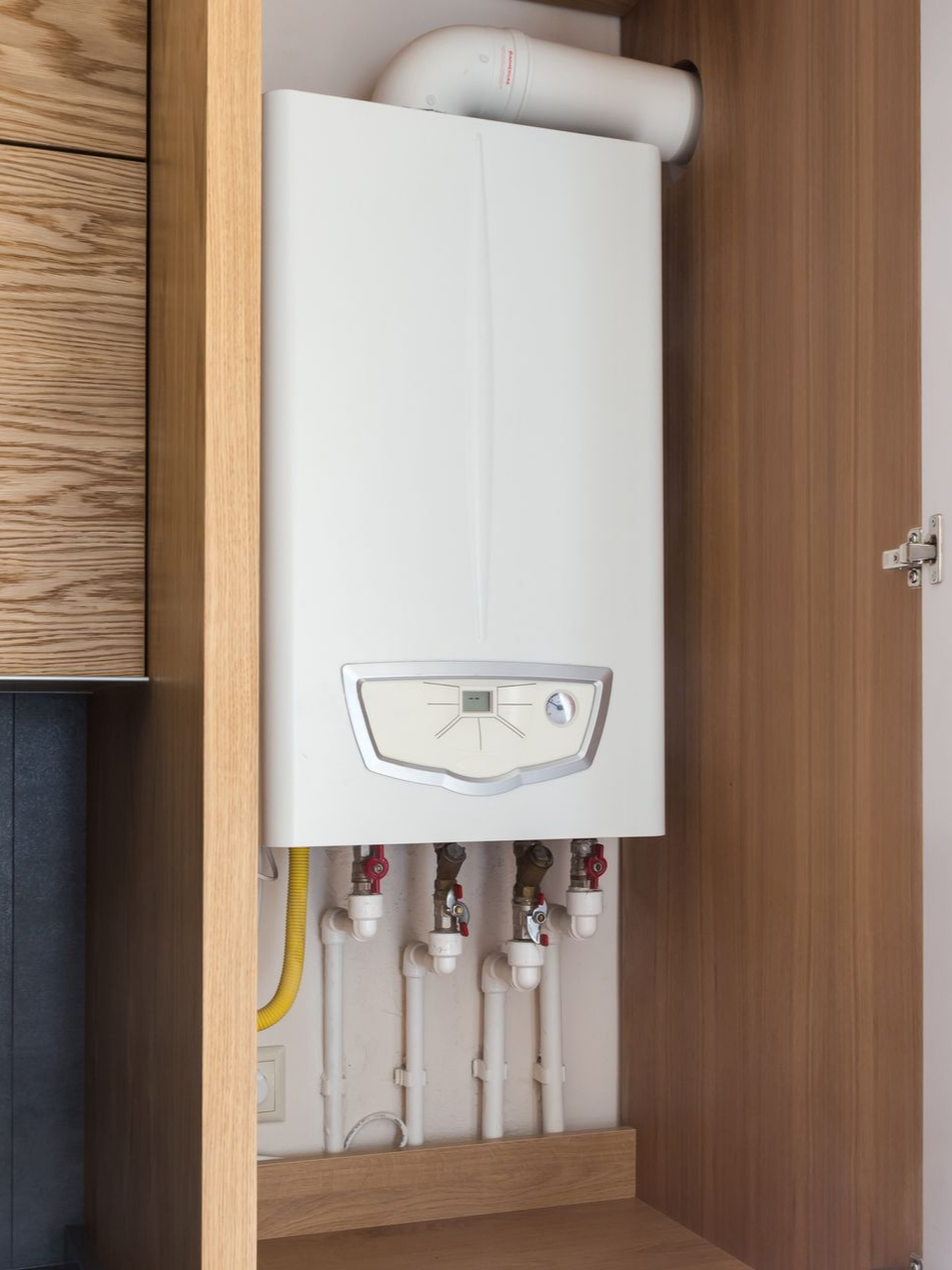 Chaudière murale blanche dans un meuble en bois, avec tuyaux et conduit d'évacuation.