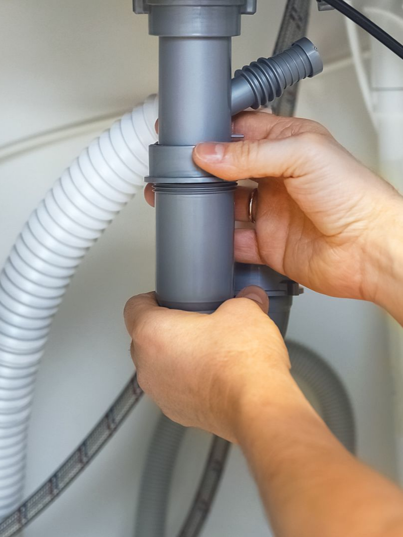 Des mains assemblent des tuyaux de plomberie en plastique gris sous un évier.