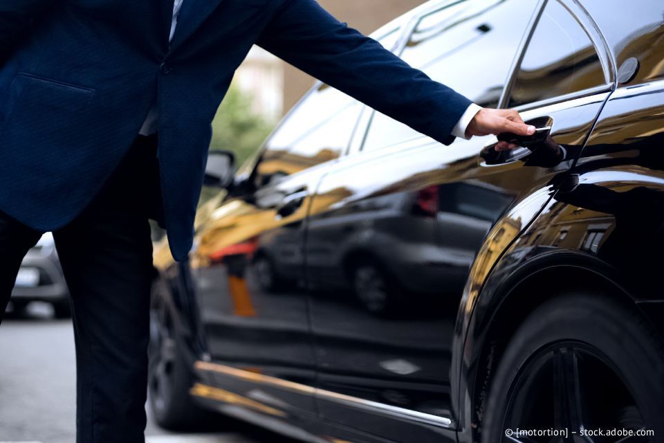 Eine Person im Anzug öffnet die Tür eines schwarzen Autos auf einer Straße in der Stadt.
