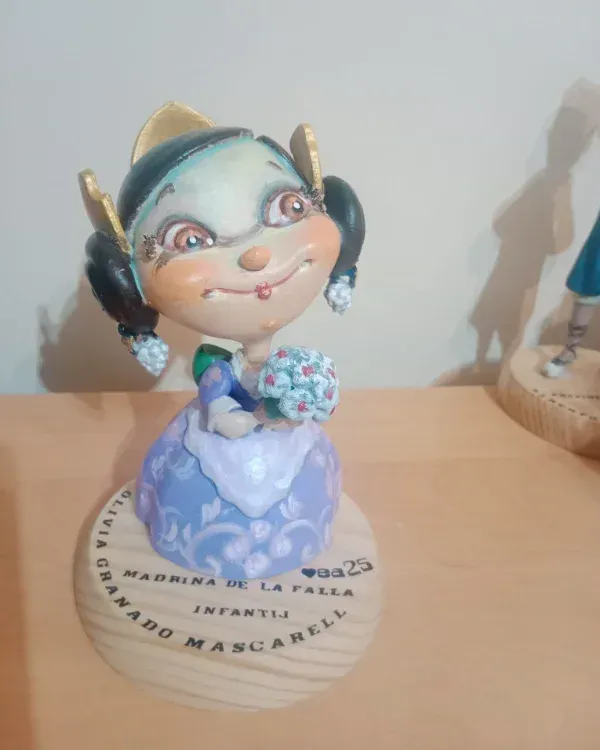Bonita figurita con un vestido azul que sostiene flores, expuesta sobre una base redonda en una mesa.