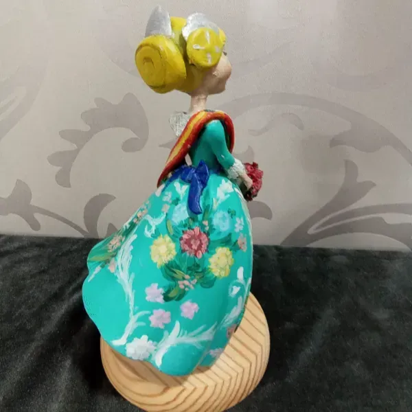Muñeca pequeña con un vestido floral turquesa sobre una base redonda de madera sobre un fondo gris estampado.