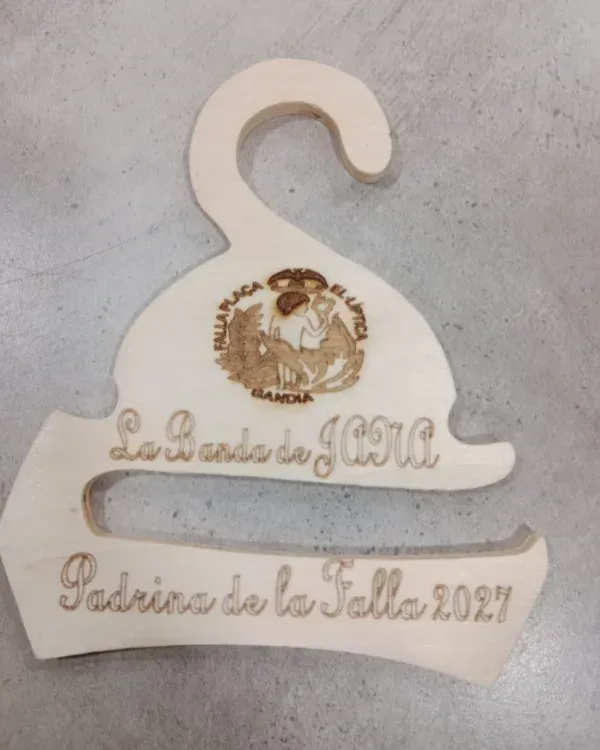 Placa blanca colgante de madera con inscripción dorada “Padrinos de la Talla 2024” sobre una superficie gris.