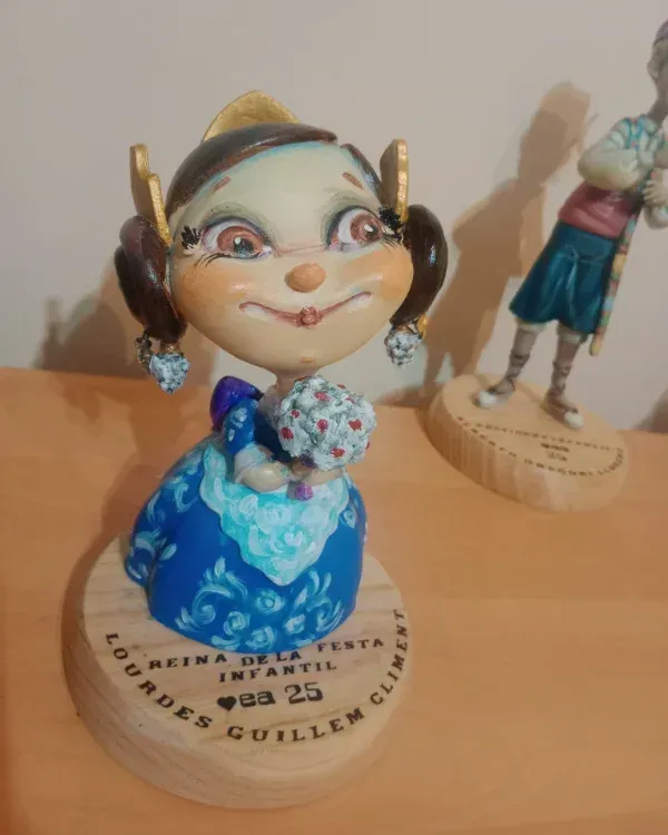 Figurita vestida de azul con elaborados accesorios para el cabello, de pie sobre una base etiquetada.