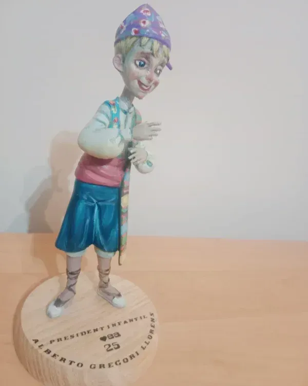 Figurita pintada de un niño sonriente con gorra azul y pantalones cortos, de pie sobre una base redonda.
