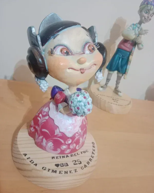 Pequeña figurita pintada de una niña sonriente con un vestido rosa que sostiene una magdalena azul.