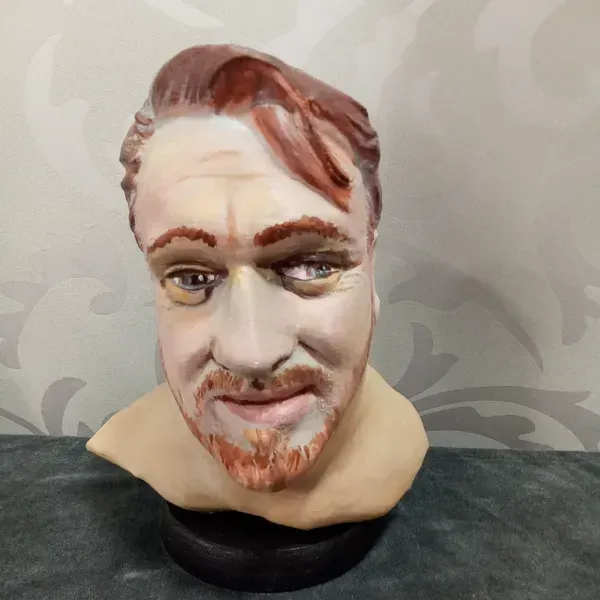 Busto de cerámica pintada con cabello castaño rojizo, ojos verdes y una leve sonrisa sobre una base negra.