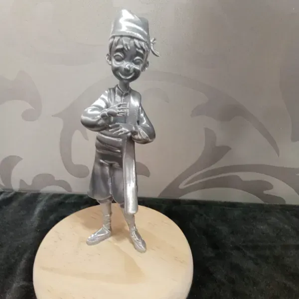 Pequeña figura plateada de dibujos animados sobre una base de madera, de pie sobre un fondo de tela oscura.