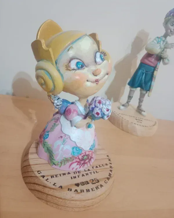 Figurita pintada a mano de una niña rubia con un vestido floral sobre una repisa de madera, con otra figurita borrosa detrás.