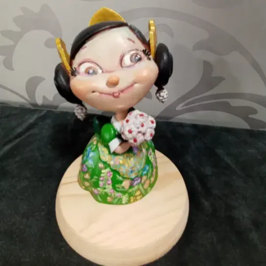 Pequeña figurita decorativa con cabello negro, vestido verde y tocado amarillo sobre una base de madera.