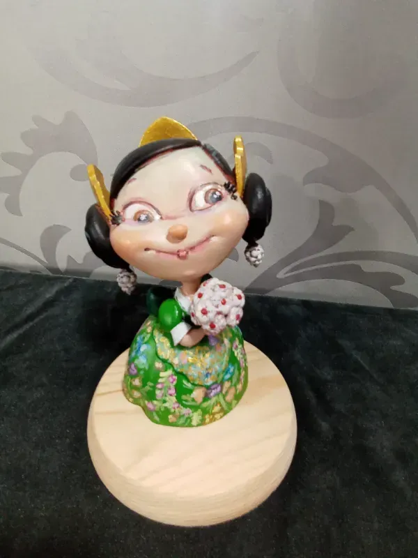 Pequeña figurita de cerámica con cabello negro y vestido verde sobre una base de madera, sosteniendo flores.