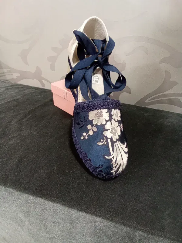 Patuco de bebé azul con estampado floral y cordones, colocado sobre una pequeña caja rosa con un fondo de tela oscura.