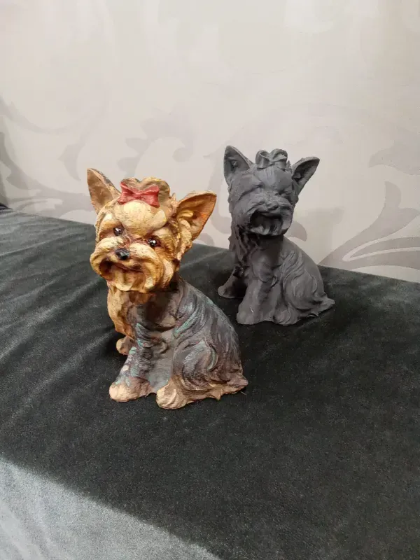 Dos pequeñas figuritas de Yorkshire terrier, una marrón y otra negra, sobre una superficie oscura.