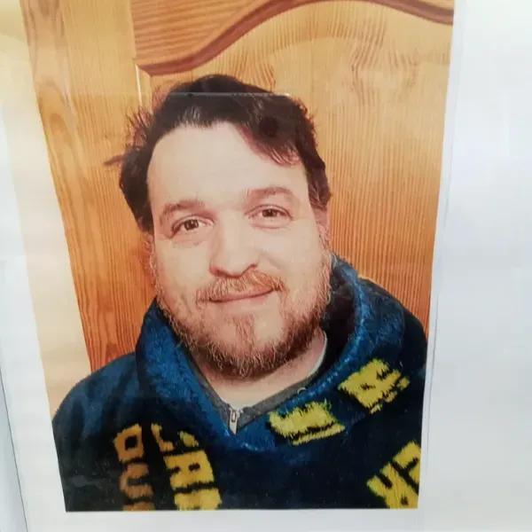 Hombre sonriente con una túnica azul, sentado contra el respaldo de una cálida silla de madera.