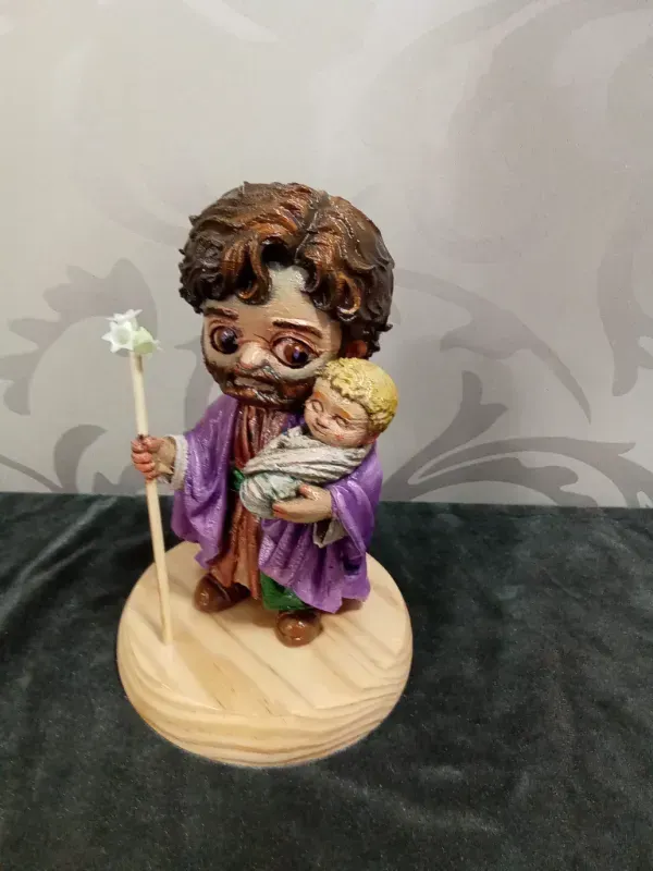 Figurilla de una figura vestida con una túnica que sostiene a un bebé, de pie sobre una base de madera con un bastón.
